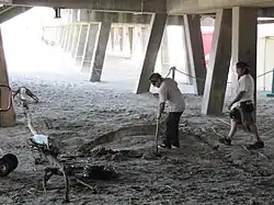 Two men, dressed in white shirts and loose pants, are seen beneath a wooden pavilion. The pavilion's legs stick out diagonally from the pavilion. One man carries a pipe which he points toward a pit in the sandy ground. The second man walks toward him.