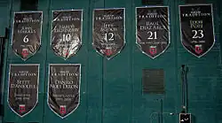 Seven large black shield-shaped banners are hung on a green wall, with white text for the name and number, or role that the individual played.