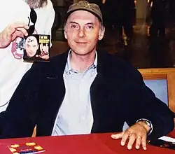A man in his forties wearing a brown cap, a light blue shirt with a very dark blue sweater. He has a slight smile, and his hand is on the red table.