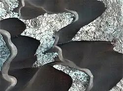 Dark sand dunes on Nili Patera of probable volcanic origin. The lighter-coloured surface is ancient lava, which has cooled. The cracks on the lava floor are filled with dark, volcanic sand.