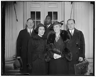 Five adults standing for a group photo: front is two white women; back row is two white men and a black man (center). They are indoors.