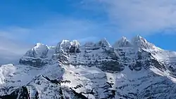 Vertical sawtooth-shaped rock walls overhanging a snowy slope lined with a few trees.