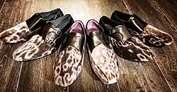 Three pairs of dress shoes on a wooden floor. The body of the shoes is covered with fur which has contrasting rings. The upper part of the shoes are shiny black leather with buckles for closure.