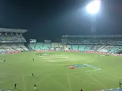 Panoramic view of a cricket ground under floodlights with players occupying the field.
