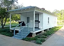 Present-day photograph of a whitewashed house, about 15 feet wide. Four banistered steps in the foreground lead up to a roofed porch that holds a swing wide enough for two. The front of the house has a door and a single-paned window. The visible side of the house, about 30 feet long, has double-paned windows.
