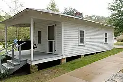 Present-day photograph of a whitewashed house, about 15&nbsp;feet wide. Four bannistered steps in the foreground lead up to a roofed porch that holds a swing wide enough for two. The front of the house has a door and a single-paned window. The visible side of the house, about 30&nbsp;feet long, has double-paned windows.