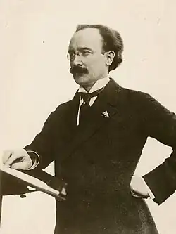 A thirtysomething man in a tuxedo and glasses clutches a violin and gazes at the camera.