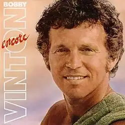 A photo of Bobby Vinton from the shoulders up smiling to his left at the camera. He is bare-chested with a green towel over his left shoulder. Beside him are the words BOBBY VINTON in white and ENCORE in red. The background is peach on the top, blue on the bottom, like a beach sunset.