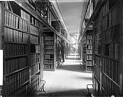 A man steps on a railing to reach a book on a high shelf, flanked by massive book shelves; a second floor is visible and in the distance is a spiral staircase