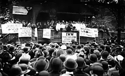 Black and white photograph of a speaker rallying a large crowd. In front of the stage, facing the audience, are several signs, in various languages, displaying demands.
