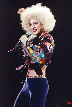A woman with short blonde hair, wearing a green bra and purple pants, singing to a microphone, held in her left hand.