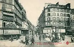 Sepia picture postcard of a commercial street scene, taken from a crossroads, looking down the street. A two-car train is in the foreground, head-on. Several pedestrians are crossing towards it.