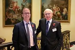 Dominic O'Connell pictured next to Dick Olver, both wearing suits and poppy appeal poppies, they are stood in front of two paintings