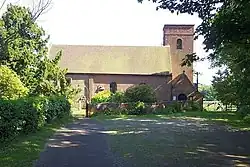 A photograph of a simple country church, with its square western tower in brick to the right and the older stone eastern end, slightly obscured by trees to the left.