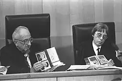 A black and white photo showing two men at a podium handling photographs of military-looking men