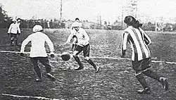 Black and white photo with three players in a white kit, and one player in a striped kit, with a football on a muddy pitch.