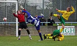 Several players standing near a goal, attempting to reach the ball. Some of them are not touching the ground, and do not appear to be completely in control of their bodies
