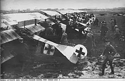 A black and white image of triplanes lined up on grass a black Iron Cross on the back of each of them with many people behind them