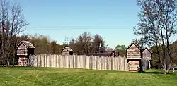 A wooden stockade fort with a green lawn in the foreground.