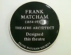 A dark green, circular plaque with the words "Frank Matcham (1854–1920) Theatre Architect Designed this theatre The Frank Matcham Society" in gold lettering.