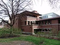 View of the house from the northern property line. The facade is made of brick with some windows. The overhanging roofs and exterior terraces are also visible.