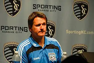A man in a blue polo shirt is sitting behind a microphone and in front of a backdrop with the Sporting Kansas City logo.