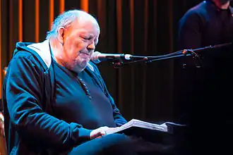 Freddie Wadling wearing a dark knit shirt and hoodie, sitting onstage in front of a microphone, looking down at a book he is holding in his lap