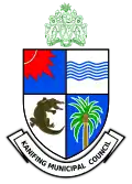 A quartered shield, blue and white. Top left has a red sun. Top right has blue waves. Bottom left has a crocodile. Bottom right has a coconut tree. Above, a green coat of arms of The Gambia. Below, a banner saying "KANIFING MUNICIPAL COUNCIL".