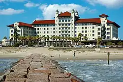 A stately white hotel building with a red-tile roof is seen from the end of a jetty extending from the beach.
