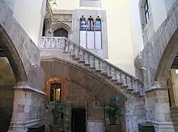 A view of a courtyard inside a building with gray stone arches to right and left and a staircase ascending from right to left across the back wall with potted plants under the stairs. Above the stairs are windows and doors of gothic design.