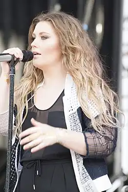 Young woman with long light coloured wavy hair wearing black clothing singing at a microphone