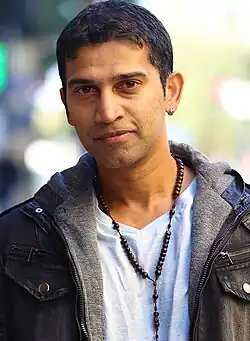 Actor Giju John standing on the set of the movie Ranam, wearing a grey jacket and white shirt