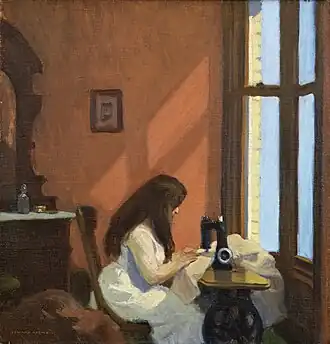 A woman sits at a sewing machine near a window in a softly lit room