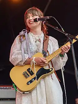 Teasdale in a white dress standing on stage with a guitar, smiling and looking to the right