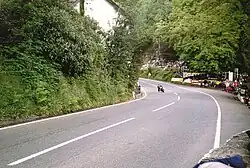 Racing motorcycle negotiating the entrance to Glen Helen turn, exiting uphill, set deep in the countryside with the entrance to the actual Glen and car park (shared with the closed restaurant) to the right, with many spectators around the outside of the bend