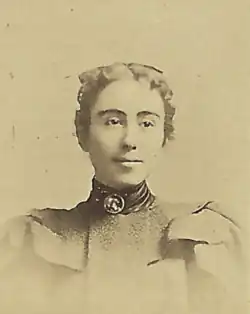 19th-century B&W portrait photo of a young woman with her hair in an up-do, wearing a dark, high-collared blouse with puffy sleeves.