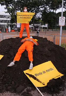 Protesters dressed in orange uniforms hold yellow signs. One of them is standing on top of a small pile of dirt and the other one is laying on it. The sign on the dirt says "pile of shit"