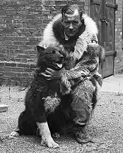 Gunnar Kaasen crouched down next to Balto, a black Siberian husky with white markings on both his chest and feet. Behind them is a brick building with a wooden door supported by wrought iron.