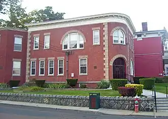 View of the library from across the street, with its retaining wall in front. It is a red brick building with some Italianate decoration and "Fowler Memorial Building" on the cream-colored frieze