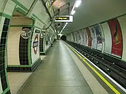 View along a platform in a circular tunnel. Tracks run on the right side with the walls covered with cream and green tiles. An illuminated 'way out' sign overhead indicates the exit.