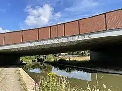 A brick and concrete road bridge spanning a canal with a dirt path on the left side, occupied by a cyclist in the distance