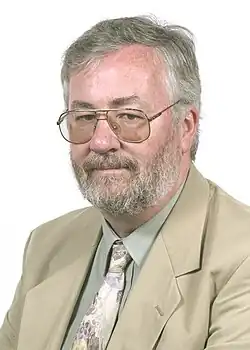 Portrait photograph of Kerr in his fifties. He is wearing a tan suit and olive shirt with a floral-patterned tie, as well as glasses. His hair and beard are greying, but retain some darker colour.