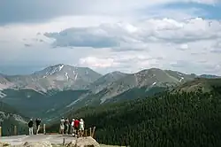 A small paved area encircled by a guardrail with wooden posts and ropes. People are standing in it looking at high snow-capped mountains in the distance
