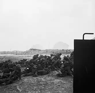 Infantry pile up on the beach. In the distance can be seen barbed wire and a gun emplacement