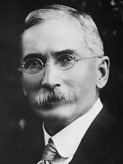 Black and white photograph of a man with a prominent mustache sporting a suit and glasses.