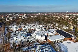 A aerial view in winter 2024 with snow on the ground and a blue sky, the John F. Kennedy School campus in the foreground and the Berlin Zehlendorf residential neighborhood with a forested park in the background