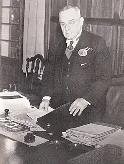 Man in a suit standing, holding a folder on a desk, while facing forward towards a camera.