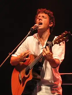 A curly haired man, strumming a guitar and wearing a white shirt.