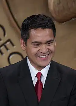 Manicad wears a suit with a red tie and is smiling while looking at something below the camera