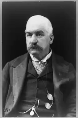 Portrait photograph of a man in a three-piece suit, with a chain (for a pocket watch?) and spectacles visible on his vest. He wears a high-collared dress shirt and is mostly balding with a thick mustache.